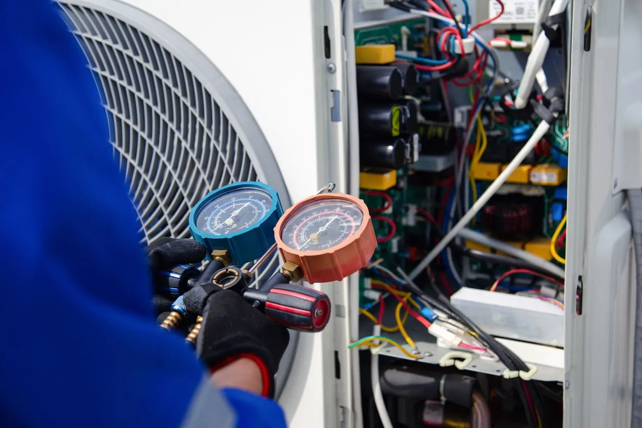 HVAC technician using gauges to inspect air conditioning unit, outdoor setting, wires visible.