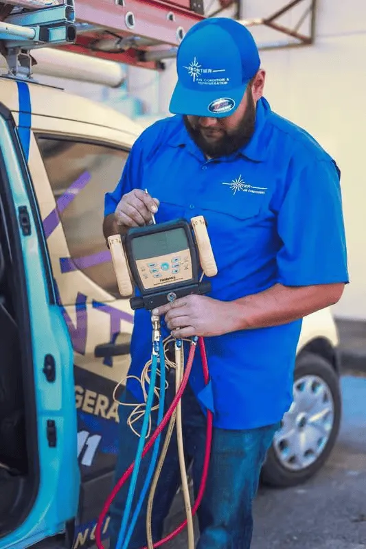 HVAC technician in blue uniform, checking gauge with hoses attached to a vehicle in an outdoor setting.